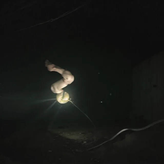 Night exterior, almost entirely black. A pair of pale bare legs folds upward out of a small glowing yellow orb resting on the ground, the body above them lost to darkness. The orb throws a hard flare across gravel; a cable snakes off to the right toward a dim shed wall.