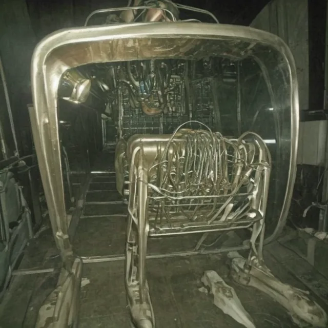 A heavy machine frame in tarnished brass and steel fills a dark industrial interior. Inside its rounded cage sits a dense basket of looping tubes and hooks, like sterilised surgical instruments. Scattered pale bones lie on the floor beneath. Sepia-green, heavily grained, part engine and part apparatus.